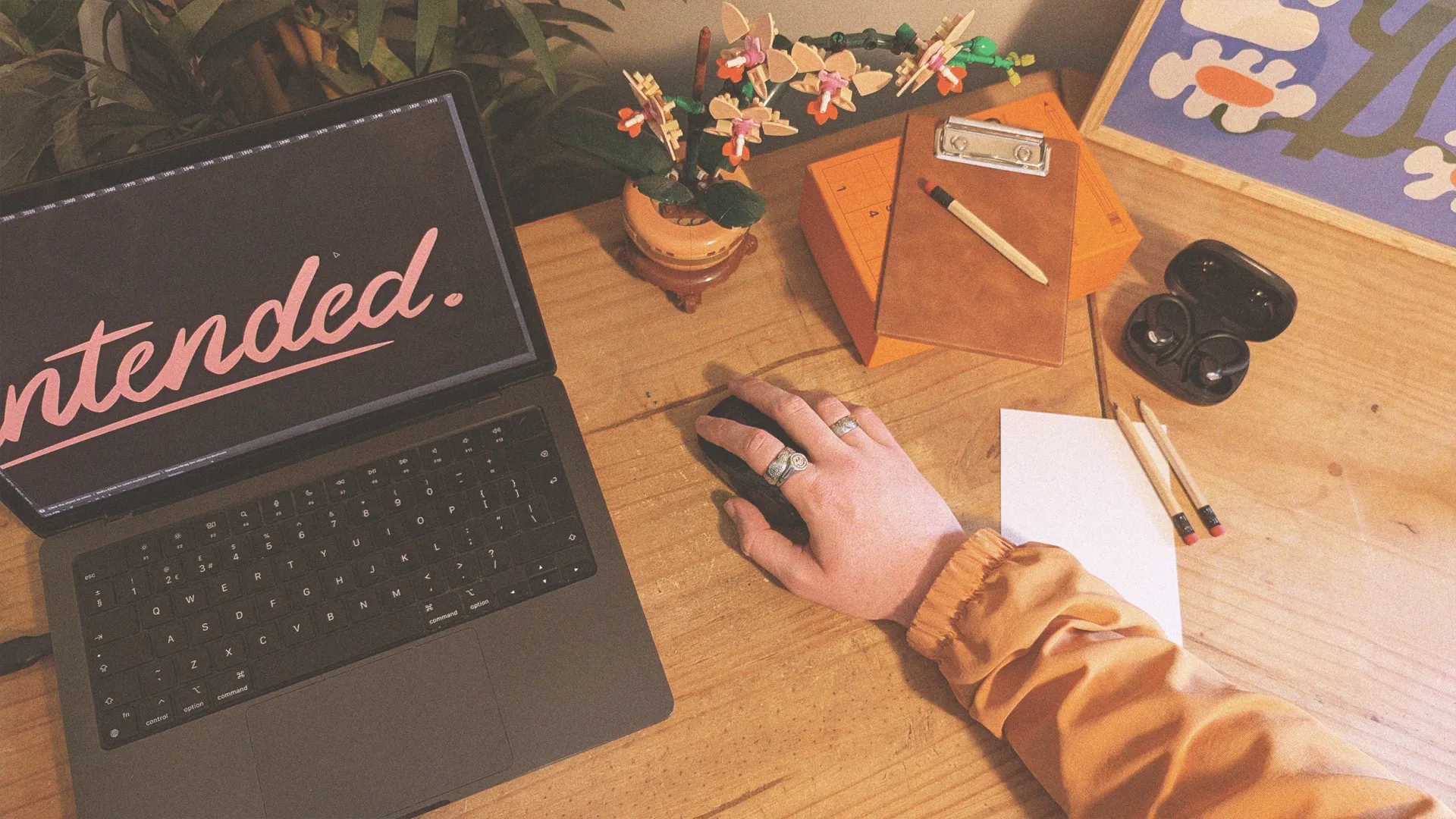 A laptop displaying the As Intended brand mark on a desk, with a hand on the mouse next to a notebook and stationery